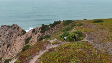Manzara, yemyeşil ve kayalık kayalıklarla çevrili bir sahil yolunda coşkulu yürüyüşçüleri gösteriyor.