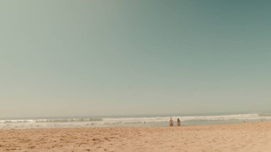 Gün batımında hafif dalgalar ayaklarına vururken altın kumda yürüyen iki figürlü sakin bir sahil sahnesi.