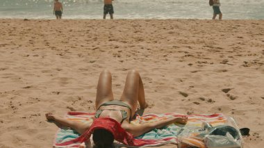 Huzurlu bir plaj sahnesinde güneşlenen biri dinlenirken, diğerleri kıyı şeridi boyunca çeşitli etkinliklerden zevk alıyor.