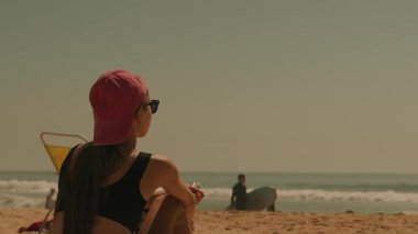 Huzurlu bir sahil sahnesi, okyanusun kenarında uzanan, sıcak güneş ışınlarının ve yumuşak sörfün tadını çıkaran bir kadını gösteriyor.