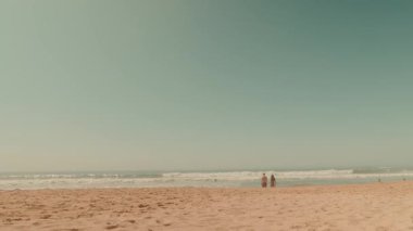Çiftler açık mavi bir gökyüzünün altında kumlu sahilde gezinirken keyifli ve huzurlu bir sahil sahnesinin tadını çıkarın.