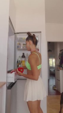 Parlak ve renkli bir mutfakta, günlük giyinmiş, mutlu bir şekilde buzdolabının yanında lezzetli yemekler hazırlayan bir kadın.