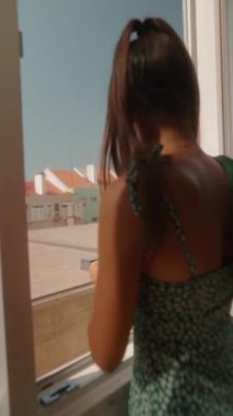 Bir kadın elinde parlak yeşil bir kumaşla pencereden dışarı bakıyor. Günlük hayatındaki huzurlu bir anı yakalıyor.