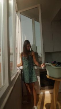 Güzel yeşil elbiseli bir kadın pencerenin yanında duruyor, sıcak güneş ışığının tadını çıkarıyor.