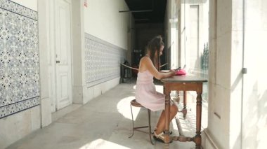 Genç bir kadın eski bir masada oturuyor, güneşli bir koridorda güzel desenli duvarları olan bir yazı yazıyor.