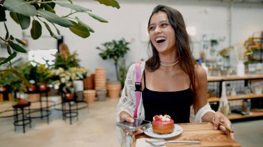 Neşeli bir kadın, yeşilliklerle çevrili canlı bir kafede pasta tutarken gülüyor.