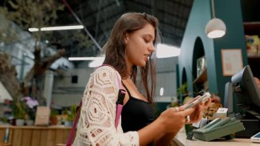 Genç bir kadın, şık ve modern bir perakende mağazasında canlı ürünlerle alışveriş yaparken aktif olarak telefonunu kullanıyor.