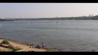 İnsanlar nehir kıyısında dinleniyor, kayıklar yüksek hızda suda yüzüyor, ve gezintiye çıkmak isteyenler.