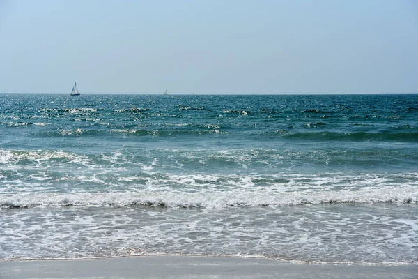 Blue ocean waves with white foam on sandy beach. Tranquil seascape with sailing boats on horizon under clear sky.