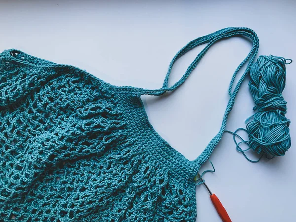 String bag in the process of knitting. Turquoise mesh bag, crochet hook and remaining yarn on a white background. The concept of creating a product.