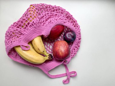 Beyaz arka planda meyveli pamuk örgü poşeti. Muzlar, elmalar, pembe iplik torbasında erikler. Sıfır israf, plastik kavram yok. Ferahlatıcı bir yaz yemeği.
