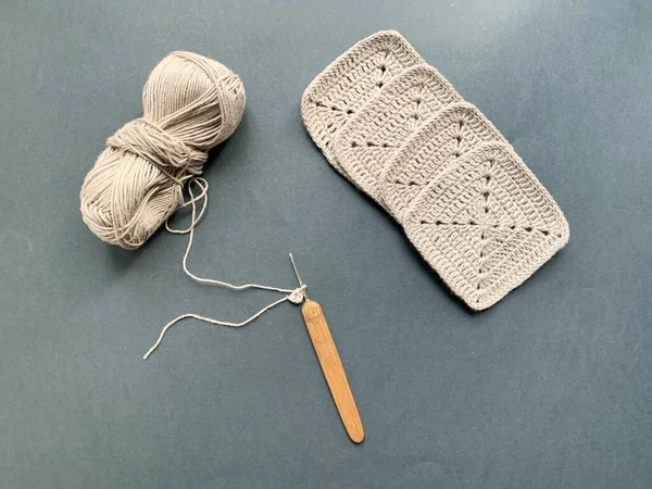 Knitting. In the photo, a hook, brown yarn, a ball of thread, knitted elements. Photo from above. The concept of creating a product. Crocheted squares on a black background.