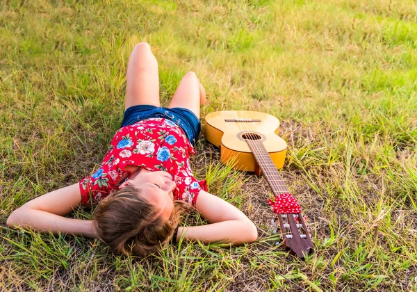 Sarışın kadın parkta çimlerin üzerinde uzanmış, yanında gitarıyla.