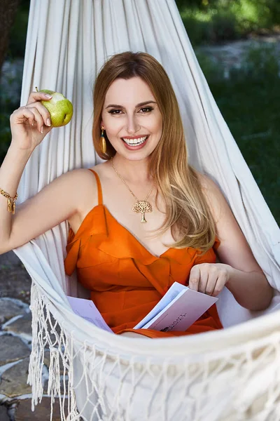 美丽的金发女模特躺在吊床上放松地呼气穿着红色丝绸衣服看书在花园里吃苹果有机食品夏天天气完美的周末 — 图库照片