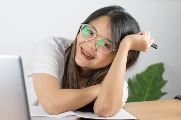 Asyalı bir kadın beyaz bir tişört giyiyor ve mutlu duygularla çalışmak için gözlük takıyor. Kameraya bakan gözleri var ve masaya dizüstü bilgisayar koyuyor ve beyaz duvar arkaplanı var.