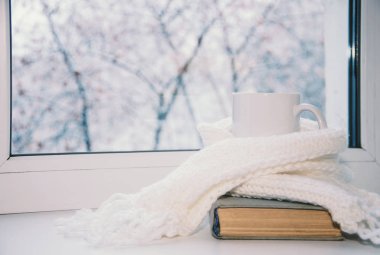 Bir kupa sıcak çay ve dışarıdaki kar manzarasına karşı cam kenarında yün örgü..