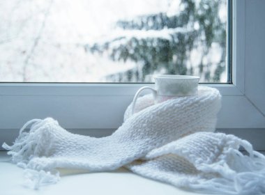 Bir kupa sıcak çay ve dışarıdaki kar manzarasına karşı cam kenarında yün örgü..