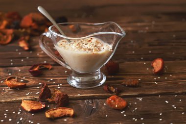 Baharatlı ekşi krema sosu ve cam bir tencerede susam tohumları, arka planda bulanık tahta bir masada. Yatay.