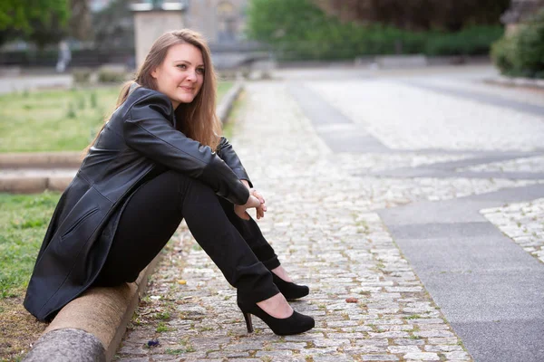 Young woman in black leather jacket sitting outdoors in park — Stock Photo, Image