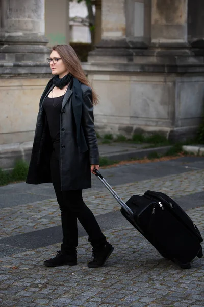 Mladá žena v černé kožené bundě chůze s kufrem — Stock fotografie