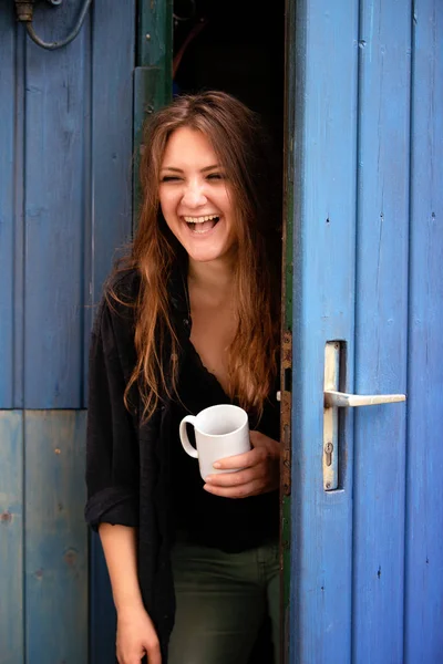 Joven morena mujer sosteniendo taza y de pie en la puerta azul —  Fotos de Stock