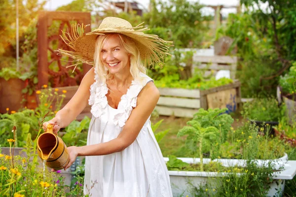 Beyaz elbiseli sarışın kadın bahçede çiçek sulama — Stok fotoğraf