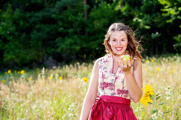 Vrouw in Dirndl staande in het veld van bloemen en het houden van een appl — Stockfoto