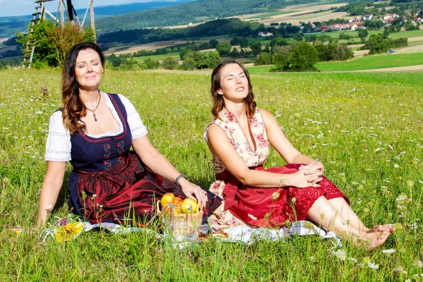 Dos mujeres en dirndl sentado en manta en el prado y disfrutando de th —  Fotos de Stock