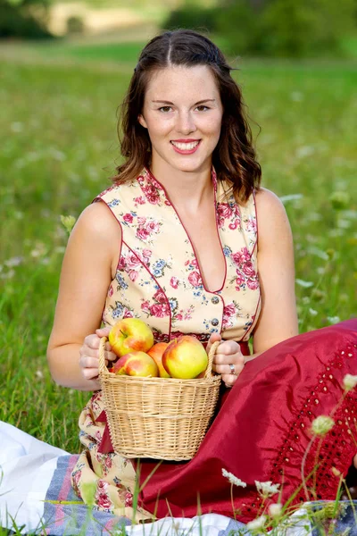 Woman in drindl sitting in a meadow and holding a basket with ap — Stock Photo, Image