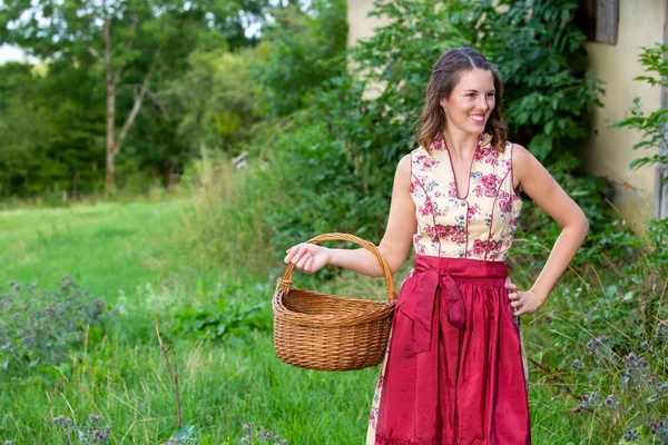 Mujer joven en dirndl de pie en la granja y la cesta de la celebración —  Fotos de Stock