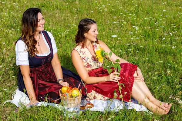 Dos mujer en dirndl sentado en manta en prado y sonriendo —  Fotos de Stock