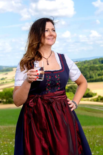 Mujer bavariana en dirndl de pie en el prado con un vaso de agua —  Fotos de Stock