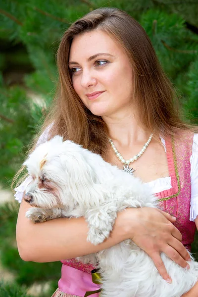 Young woman in dirndl standing outdoors and holding cute white d — Stock Photo, Image