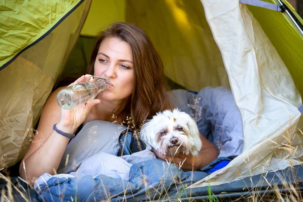 Junge Frau liegt mit Hund im Zelt und trinkt Wasser — Stockfoto