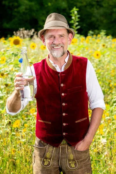 Hombre bavariano de pie en el campo de los girasoles y sosteniendo una botella —  Fotos de Stock