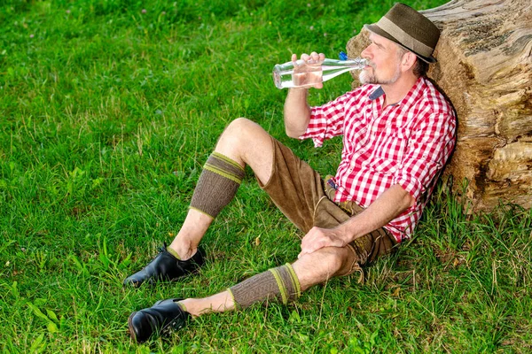 Handsome bavarian man sitting outdoors at tree stump and drinkin — Stock Photo, Image