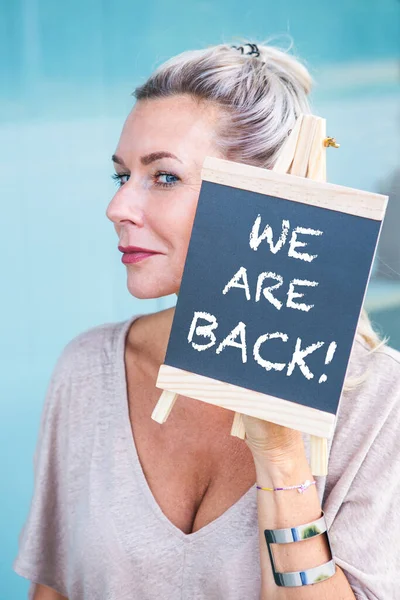 blond woman holding a chalkboard with the words We Are Back