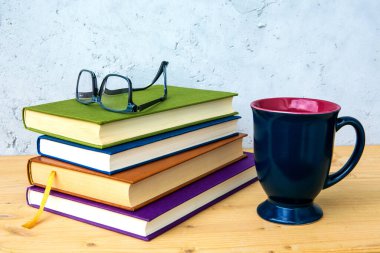 Ahşap masanın üzerinde gözlüklü bir yığın kitap ve gri arka planda bir fincan.
