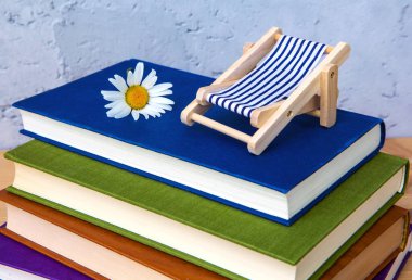 Bir yığın kitap, minyatür bir şezlong ve gri arka planı olan bir papatya.