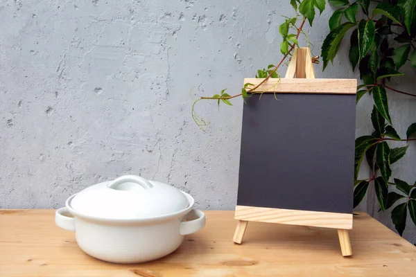 灰色の背景を持つ木製のテーブルの上に白い鍋付きの小さな黒板 — ストック写真