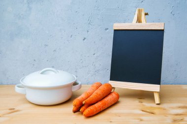 Tahtada küçük bir karatahta üzerinde havuç ve gri arka planda beyaz tencere var.