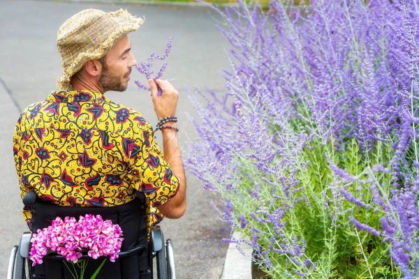 坐在轮椅上的英俊男子在户外嗅出紫色的花朵 — 图库照片