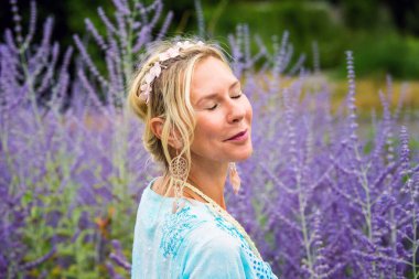 40 'larında sarışın bir kadın mor çiçeklerle dolu bir tarlada gözleri kapalı duruyor.