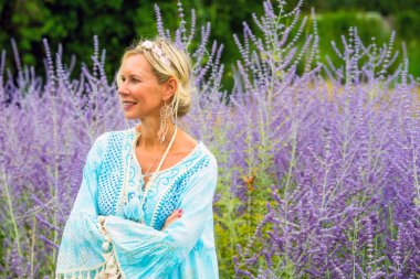 40 'larında güzel sarışın bir kadın mor çiçeklerle dolu bir tarlada dikiliyor.