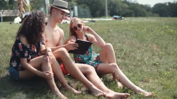 朋友们坐在草地上看着他们的平板电脑 快乐混合族族群的朋友使用电脑片剂 — 图库视频影像