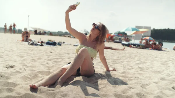 Pretty woman in bikini sitting on the beach and taking a selfie.