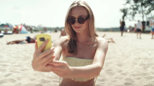 Pretty woman in bikini sitting on the beach and taking a selfie.