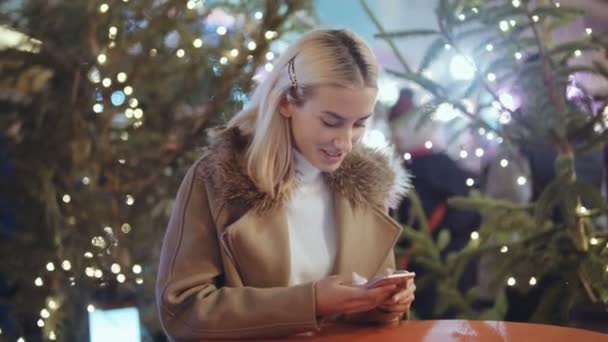 クリスマス マーケット上のスマート フォンを使用しての女性 冬を楽しんでいる女の子のホリデー シーズン ソーシャルネットワー キング 背景に約ぼやけてクリスマス ライトを使用しての概念 — ストック動画