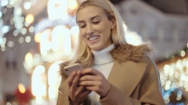 Noel pazarı üzerinde akıllı telefon kullanan kadın. Kız kış zevk tatil sezonu. İletişim, arka planda uygulama bulanık Noel ışıkları kullanarak sosyal ağ kavramı.