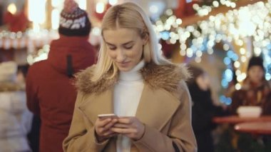 Noel pazarı üzerinde akıllı telefon kullanan kadın. Kız kış zevk tatil sezonu. İletişim, arka planda uygulama bulanık Noel ışıkları kullanarak sosyal ağ kavramı.